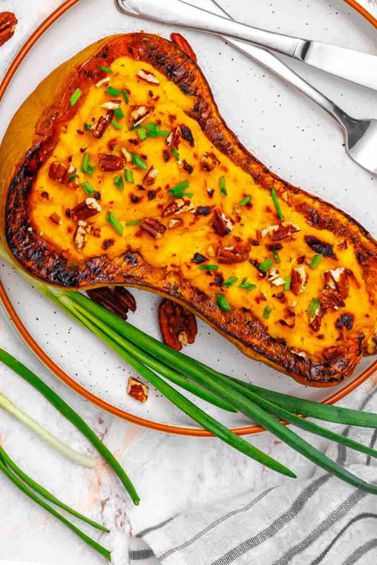 Half of a baked butternut squash filled with mashed squash, topped with chopped pecans and herbs, on a plate with green onions and cutlery.