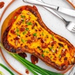 Roasted half butternut squash filled with mashed squash, topped with chopped pecans and herbs on a white plate with green onions, fork, and knife.