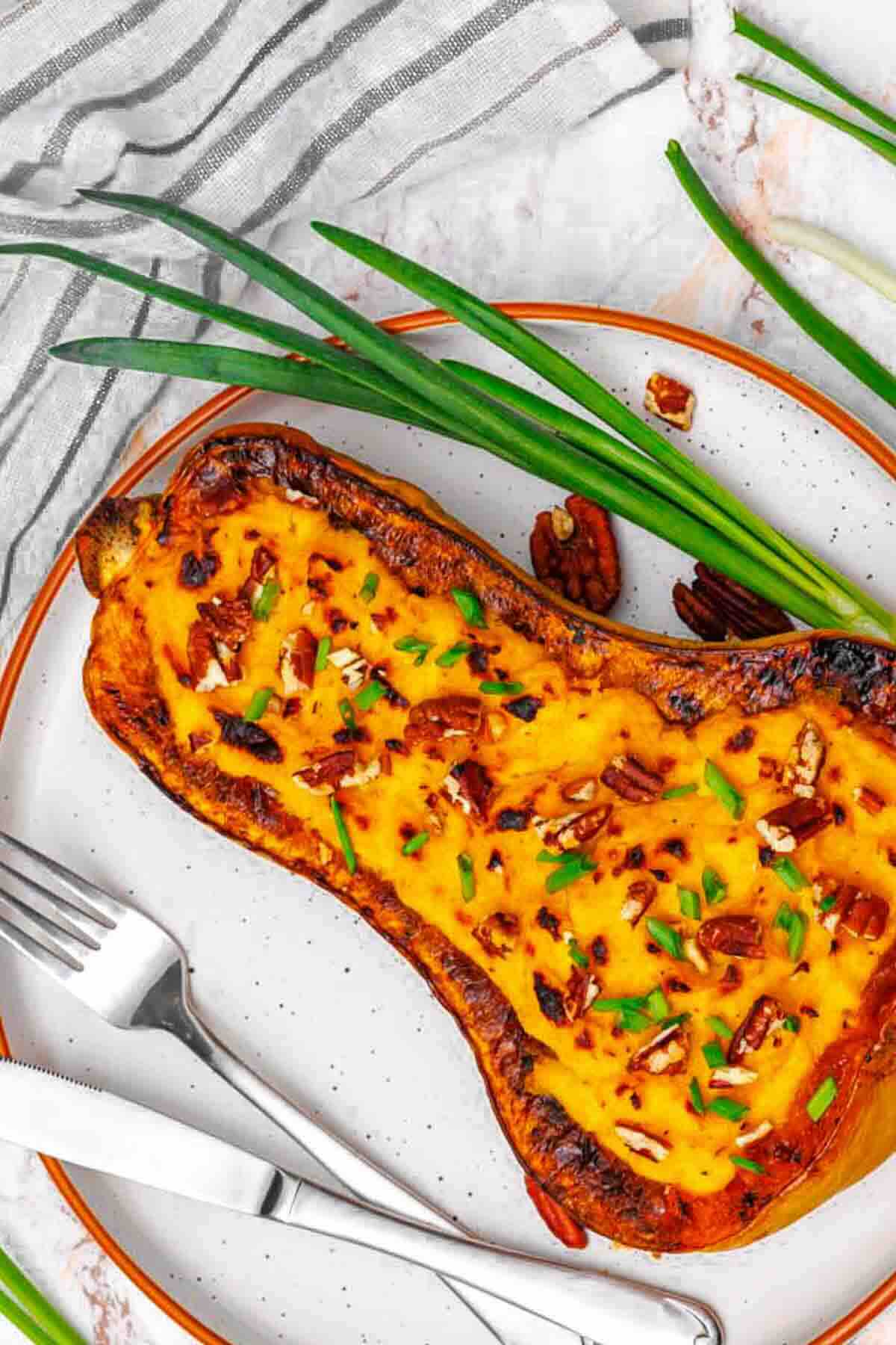 Halved roasted butternut squash filled with a cheesy mixture, topped with chopped pecans and herbs, served on a plate with fork, knife, and green onions.