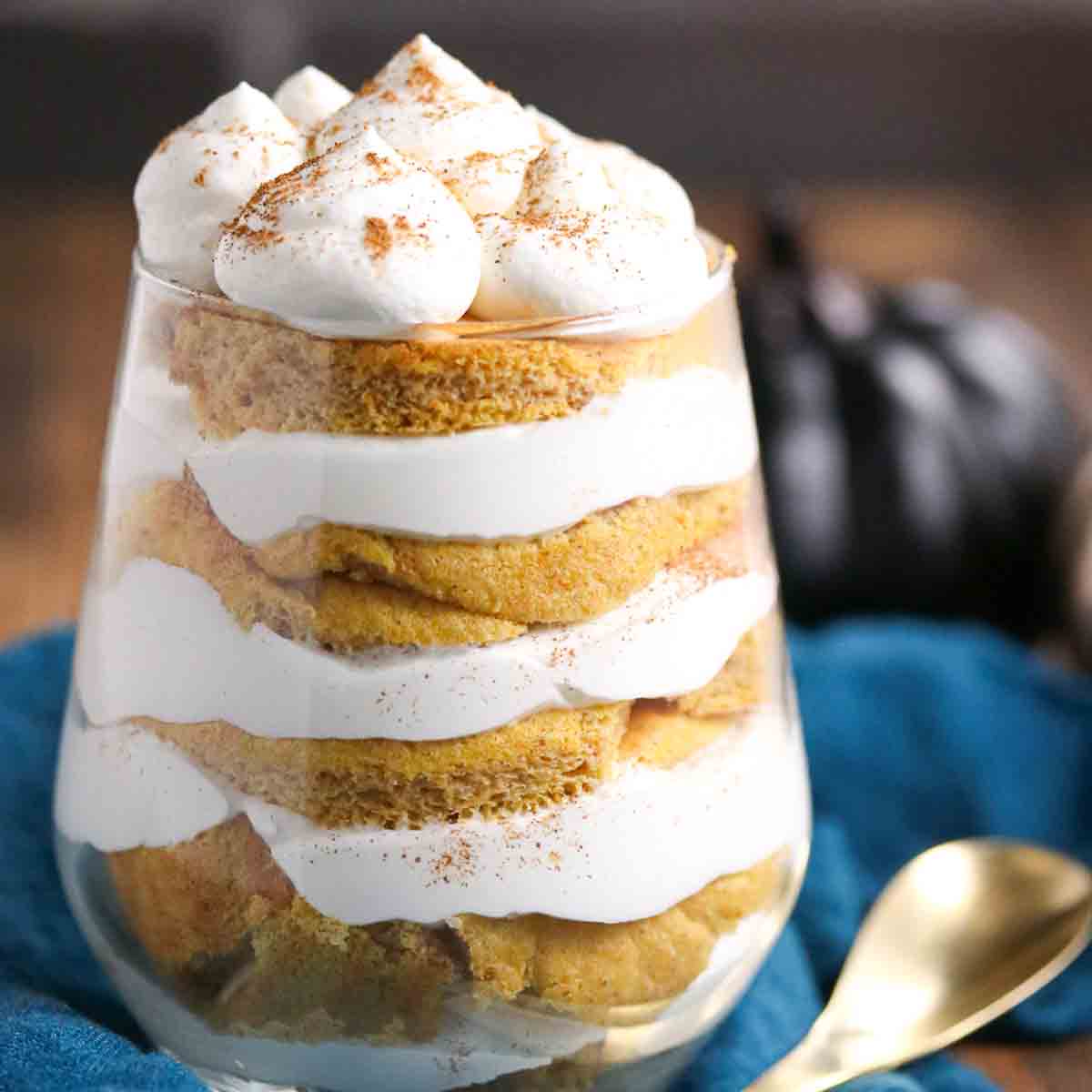 A glass filled with alternating layers of cream and cake, topped with dollops of whipped cream and a dusting of cinnamon, sits on a table next to a gold spoon.