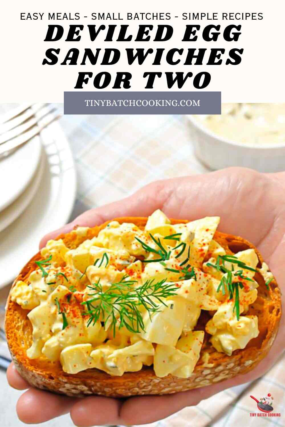 A hand holds a slice of toasted bread topped with deviled egg salad, garnished with dill and paprika. Text reads: “Deviled Egg Sandwiches for Two, tinybatchcooking.com.”