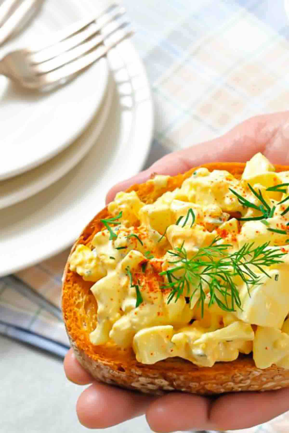 A hand holds a slice of toasted bread topped with egg salad, garnished with paprika and fresh dill.
