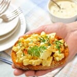A hand holding a slice of toasted bread topped with egg salad, garnished with fresh dill and paprika.