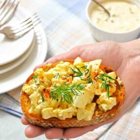 A hand holding a slice of toasted bread topped with egg salad, garnished with fresh dill and paprika.