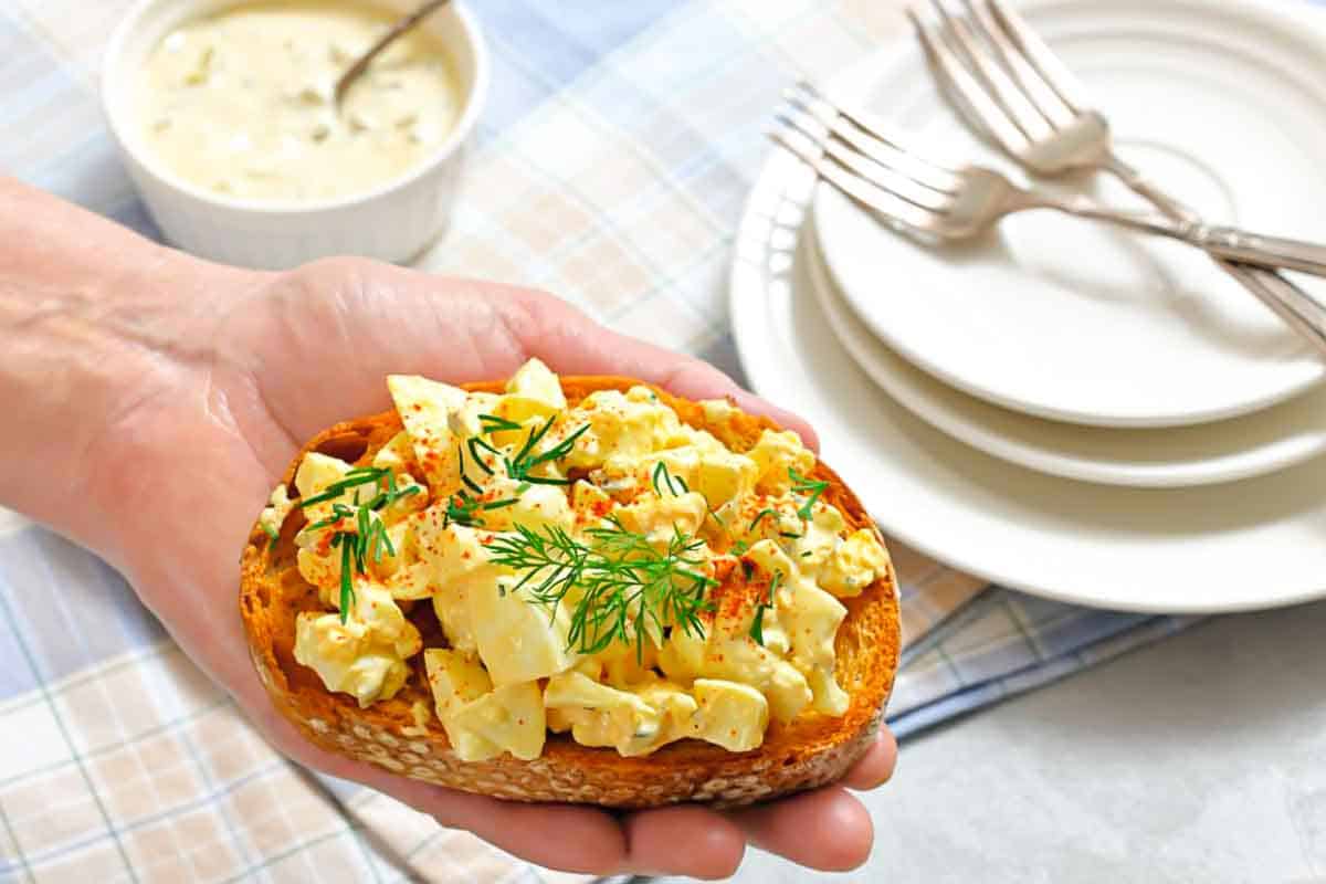 A hand holding a slice of toasted bread topped with egg salad, garnished with dill and paprika.