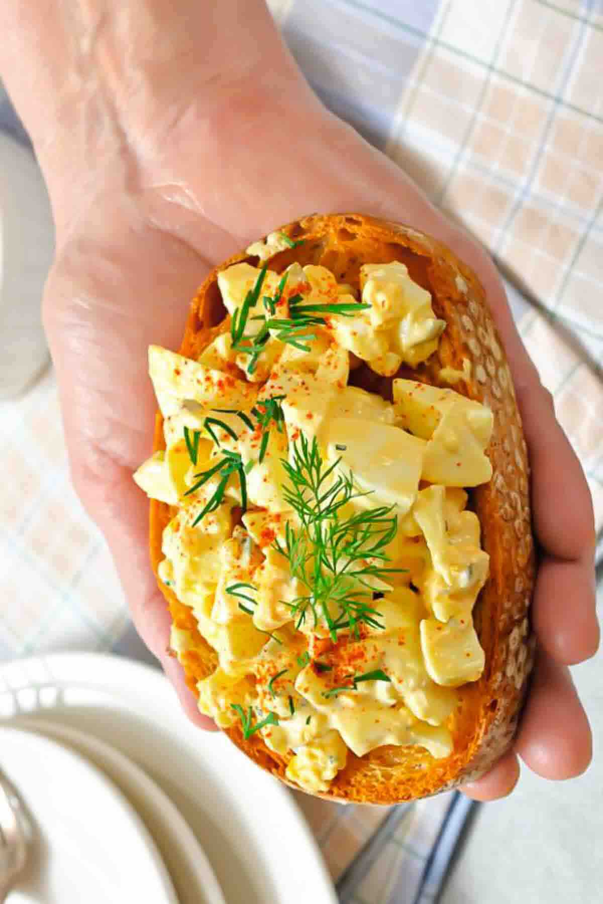 A hand holding a slice of toasted bread topped with egg salad, garnished with dill and paprika.