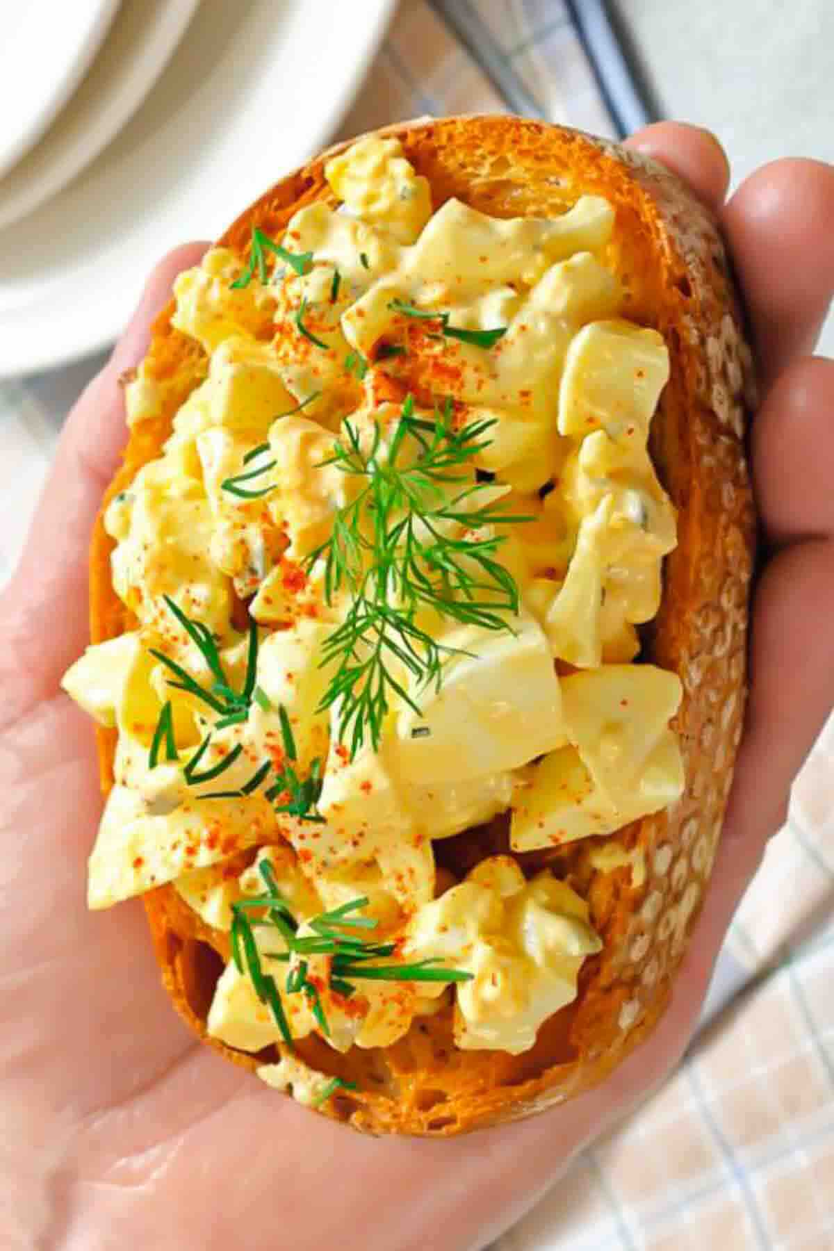 A hand holding a slice of toasted bread topped with egg salad, garnished with dill and paprika.