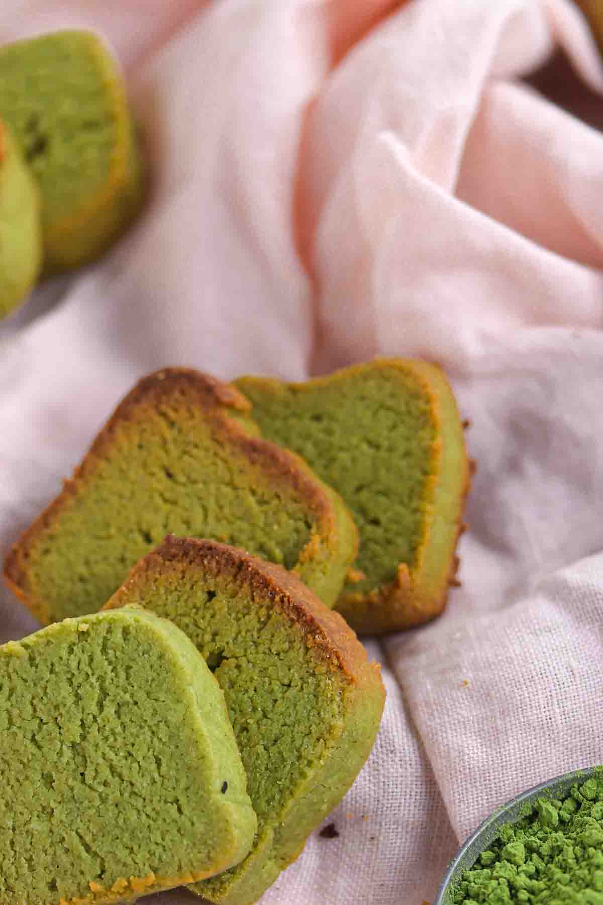 Slices of green matcha pound cake on a pink cloth, next to a small bowl of matcha powder.