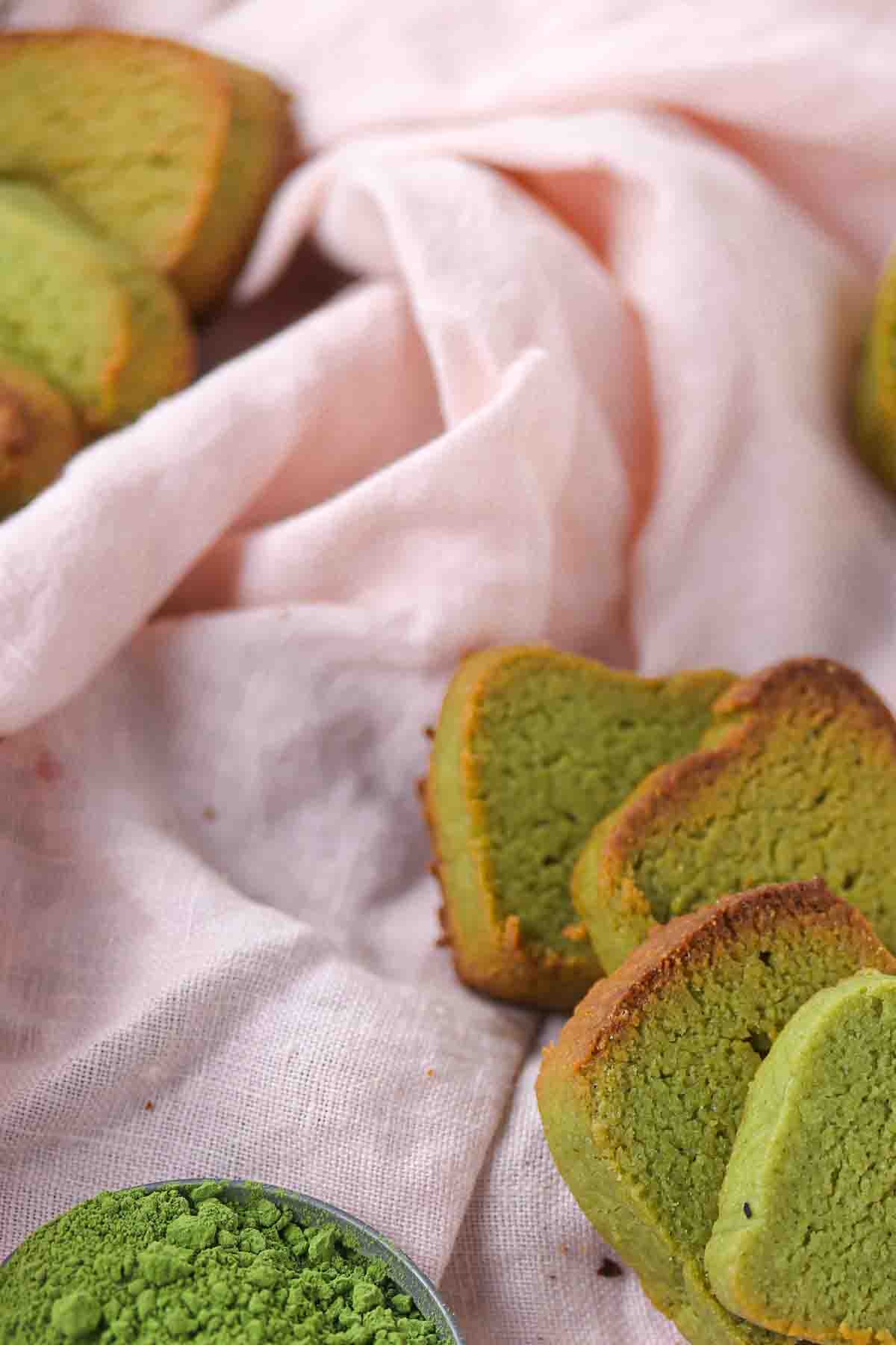 Slices of green matcha cake arranged near a small bowl of matcha powder on a light-colored cloth.