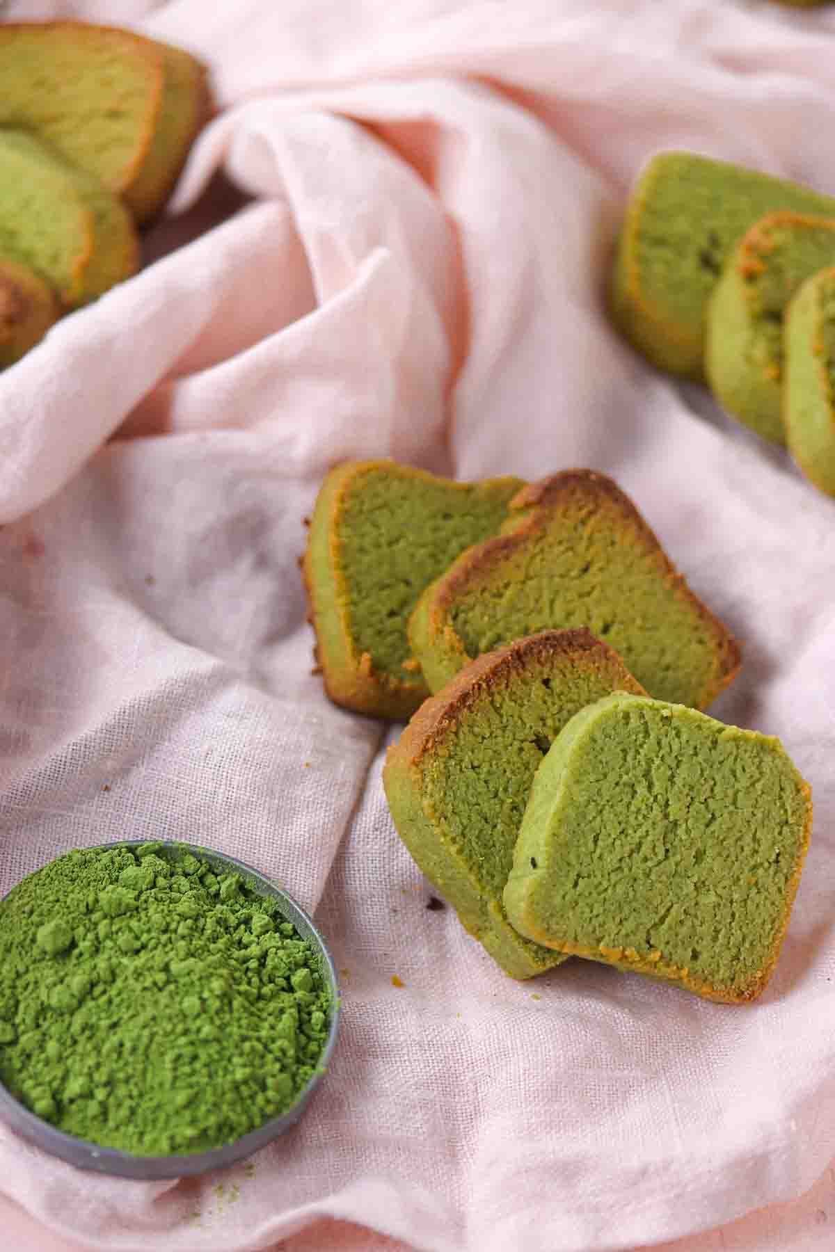Slices of green matcha cake arranged on a pink cloth with a small bowl of matcha powder.